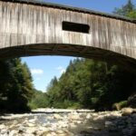 Eine überdachte Holzbrücke führt über ein felsiges Flussbett, umgeben von grünen Bäumen und unter einem klaren, blauen Himmel.