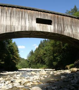 Eine überdachte Holzbrücke führt über ein felsiges Flussbett, umgeben von grünen Bäumen und unter einem klaren, blauen Himmel.