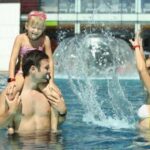 Ein Mann trägt ein lächelndes junges Mädchen auf seinen Schultern in einem Pool, während eine Frau in der Nähe Wasser spritzt.