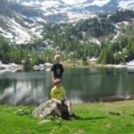 Zwei Kinder befinden sich im Freien an einem See, wobei eines auf einem Felsen sitzt und das andere dahinter steht, umgeben von Gras, Bäumen und schneebedeckten Bergen im Hintergrund.