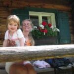 Ein Erwachsener und ein kleines Kind sitzen auf einer Holzbank vor einer rustikalen Hütte mit einem Blumenkasten unter einem Fenster.