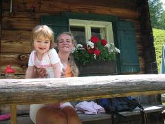 Ein Erwachsener und ein kleines Kind sitzen auf einer Holzbank vor einer rustikalen Hütte mit einem Blumenkasten unter einem Fenster.