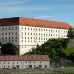 Großes rechteckiges historisches Gebäude mit rotem Ziegeldach, weißem Äußeren und vielen Fenstern, gelegen auf einem Hügel mit Grünflächen und einem kleineren Gebäude im Vordergrund.