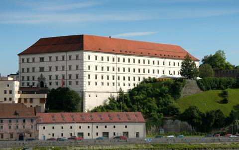 Großes rechteckiges historisches Gebäude mit rotem Ziegeldach, weißem Äußeren und vielen Fenstern, gelegen auf einem Hügel mit Grünflächen und einem kleineren Gebäude im Vordergrund.