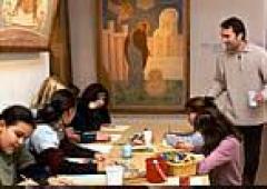 Eine Gruppe von Menschen sitzt an einem Tisch und arbeitet an Kunstprojekten, während ein Mann mit einer Tasse in der Hand in einem mit Gemälden dekorierten Raum steht.