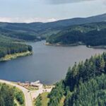 Luftaufnahme eines Flusses oder Stausees, umgeben von dichtem grünem Wald und Hügeln, mit einem kleinen Gebäude und einer Straße in der Nähe des Ufers.