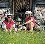 Drei Personen mit Helmen sitzen mit Fahrrädern im Gras vor einem Gebäude aus Stein und Holz.