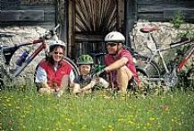 Drei Personen mit Helmen sitzen mit Fahrrädern im Gras vor einem Gebäude aus Stein und Holz.