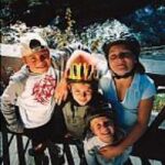 Vier Kinder mit Helmen und Freizeitkleidung stehen eng beieinander und lächeln in die Kamera auf einer Holzterrasse im Freien vor einem malerischen Hintergrund.