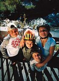 Vier Kinder mit Helmen und Freizeitkleidung stehen eng beieinander und lächeln in die Kamera auf einer Holzterrasse im Freien vor einem malerischen Hintergrund.