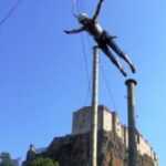 Eine Person, die einen Helm und ein Gurtzeug trägt, schwingt zwischen zwei hohen Stangen mit einer Burg auf einem Hügel im Hintergrund unter einem klaren blauen Himmel.