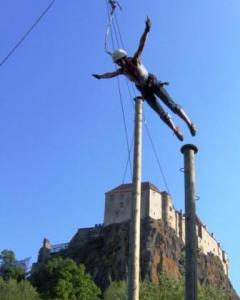 Eine Person, die einen Helm und ein Gurtzeug trägt, schwingt zwischen zwei hohen Stangen mit einer Burg auf einem Hügel im Hintergrund unter einem klaren blauen Himmel.