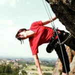 Person im roten Hemd beim Klettern im Freien, gesichert mit einem Gurt und einem Seil, mit einer ländlichen Landschaft im Hintergrund.