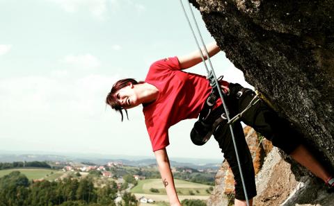 Person im roten Hemd beim Klettern im Freien, gesichert mit einem Gurt und einem Seil, mit einer ländlichen Landschaft im Hintergrund.