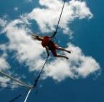 Eine Person in roter Kleidung schwebt an Bungee-Seilen in der Luft vor einem blauen Himmel mit vereinzelten Wolken.