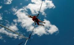 Eine Person in roter Kleidung schwebt an Bungee-Seilen in der Luft vor einem blauen Himmel mit vereinzelten Wolken.