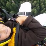 Person mit gelbem Helm und Sicherheitsausrüstung beim Gleitschirmfliegen über einem Wald, von oben fotografiert, mit Bäumen im Hintergrund.