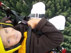 Person mit gelbem Helm und Sicherheitsausrüstung beim Gleitschirmfliegen über einem Wald, von oben fotografiert, mit Bäumen im Hintergrund.