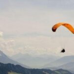 Eine Person, die mit einem orangefarbenen Fallschirm über eine Gebirgslandschaft bei teilweise bewölktem Himmel gleitet.