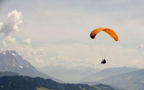 Eine Person, die mit einem orangefarbenen Fallschirm über eine Gebirgslandschaft bei teilweise bewölktem Himmel gleitet.
