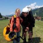 Ein junges Mädchen mit Helm und Ausrüstung steht neben einem Erwachsenen mit Gleitschirmausrüstung auf einer Wiese mit Hügeln und Häusern im Hintergrund.