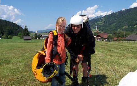 Ein junges Mädchen mit Helm und Ausrüstung steht neben einem Erwachsenen mit Gleitschirmausrüstung auf einer Wiese mit Hügeln und Häusern im Hintergrund.