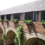 Eine gemauerte Arkade mit Bögen und einem überdachten Gang darüber, mit Säulen und Grünpflanzen auf der oberen Ebene.