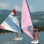 Zwei Personen beim Windsurfen auf einem See, jede auf einem eigenen Brett mit bunten Segeln, mit Hügeln und Bäumen im Hintergrund.