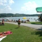 An einem sonnigen Tag entspannen sich die Menschen im Gras und schwimmen an einem Strand am Seeufer, wobei Sonnenschirme und eine Rutsche in der Nähe des Wassers zu sehen sind.