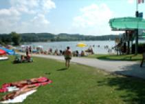 An einem sonnigen Tag entspannen sich die Menschen im Gras und schwimmen an einem Strand am Seeufer, wobei Sonnenschirme und eine Rutsche in der Nähe des Wassers zu sehen sind.