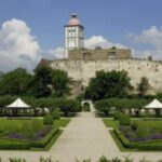 Ein historisches Steinschloss mit einem weißen Turm steht auf einem Hügel, umgeben von formalen Gärten, Bäumen und Wanderwegen unter einem teilweise bewölkten Himmel.