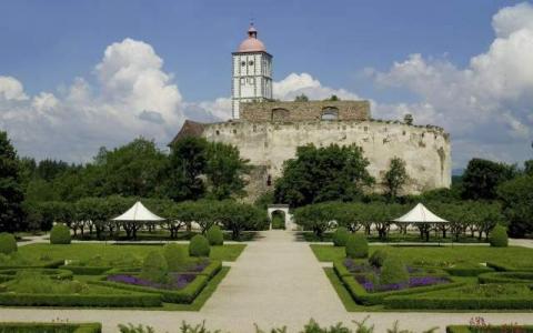 Ein historisches Steinschloss mit einem weißen Turm steht auf einem Hügel, umgeben von formalen Gärten, Bäumen und Wanderwegen unter einem teilweise bewölkten Himmel.
