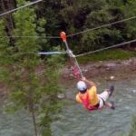 Eine Person, die einen Helm und ein Gurtzeug trägt, fährt mit einer Seilrutsche über einen Fluss, mit Bäumen und Grünflächen im Hintergrund.