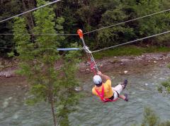 Eine Person, die einen Helm und ein Gurtzeug trägt, fährt mit einer Seilrutsche über einen Fluss, mit Bäumen und Grünflächen im Hintergrund.