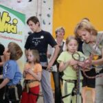 Erwachsene und Kinder mit Klettergurten stehen in einer Gruppe in einer Halle und hantieren mit Kletterseilen, im Hintergrund ist ein Banner von Wiki Kinderbetreuung zu sehen.