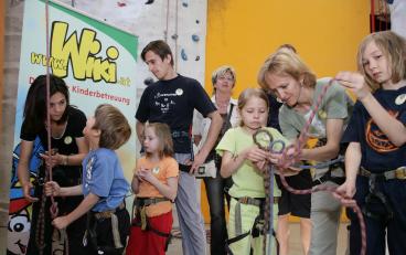 Erwachsene und Kinder mit Klettergurten stehen in einer Gruppe in einer Halle und hantieren mit Kletterseilen, im Hintergrund ist ein Banner von Wiki Kinderbetreuung zu sehen.