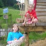 Zwei Kinder spielen auf einer Holzkonstruktion im Freien; ein Mädchen steht auf dem Boden, während das andere auf der Plattform kniet. Im Hintergrund sind ein blaues Schild und Grünpflanzen zu sehen.