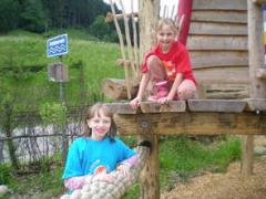 Zwei Kinder spielen auf einer Holzkonstruktion im Freien; ein Mädchen steht auf dem Boden, während das andere auf der Plattform kniet. Im Hintergrund sind ein blaues Schild und Grünpflanzen zu sehen.