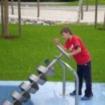 Ein Junge in einem roten Hemd dreht eine archimedische Schraube aus Metall in einem Außenbereich mit Gras und Bäumen.