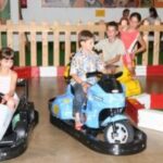 Vier Kinder fahren im Haus Autoscooter, zwei in Autos und zwei auf Motorrädern, umgeben von einem Holzzaun und rot-weißen Absperrungen.