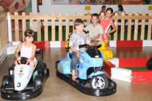 Vier Kinder fahren im Haus Autoscooter, zwei in Autos und zwei auf Motorrädern, umgeben von einem Holzzaun und rot-weißen Absperrungen.