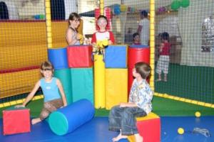 Vier Kinder spielen mit großen Schaumstoffblöcken in einem mit Netzen eingefassten Innenspielbereich. Andere Kinder sind im Hintergrund zu sehen.