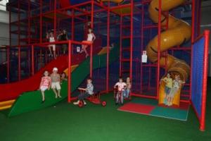 Kinder spielen auf Rutschen, Klettergerüsten und fahren auf einem Dreirad in einem Indoor-Spielplatz mit bunten Geräten und gepolsterten Böden.