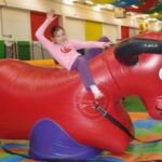 Ein Kind reitet auf einem großen roten aufblasbaren Stier in einem bunten Indoor-Spielbereich, hebt einen Arm und lächelt.