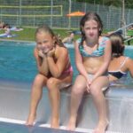 Zwei junge Mädchen in Badeanzügen sitzen an einem sonnigen Tag am Rande eines Freibads, während im Hintergrund andere Kinder schwimmen und spielen.