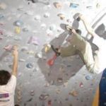 Zwei Personen klettern in der Halle an einer künstlichen Kletterwand, wobei eine Person höher klettert, während die andere nach den Griffen unten greift.