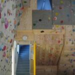 Indoor-Kletterhalle mit Wänden, die mit bunten Klettergriffen bedeckt sind, einer gelben Tür, einer Uhr und einer Treppe, die hinter der Wand nach oben führt.
