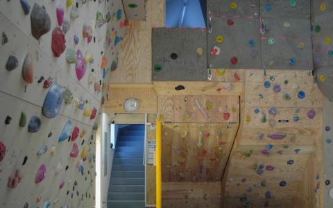 Indoor-Kletterhalle mit Wänden, die mit bunten Klettergriffen bedeckt sind, einer gelben Tür, einer Uhr und einer Treppe, die hinter der Wand nach oben führt.
