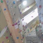 Indoor-Kletterwand mit verschiedenfarbigen Griffen und Kletterrouten, von unten betrachtet, in einem hohen, gut beleuchteten Raum.