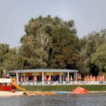 Ein Strand am See mit einem Spielplatz, einem Gebäude mit Sitzgelegenheiten im Freien und Reihen von orangefarbenen Sonnenschirmen, umgeben von Bäumen.